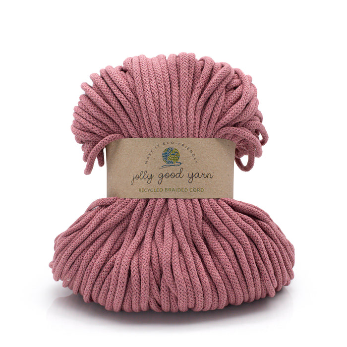 A skein of dusty rose recycled braided cord yarn by Jolly Good Yarn, wrapped in a kraft paper label, on a white background.
